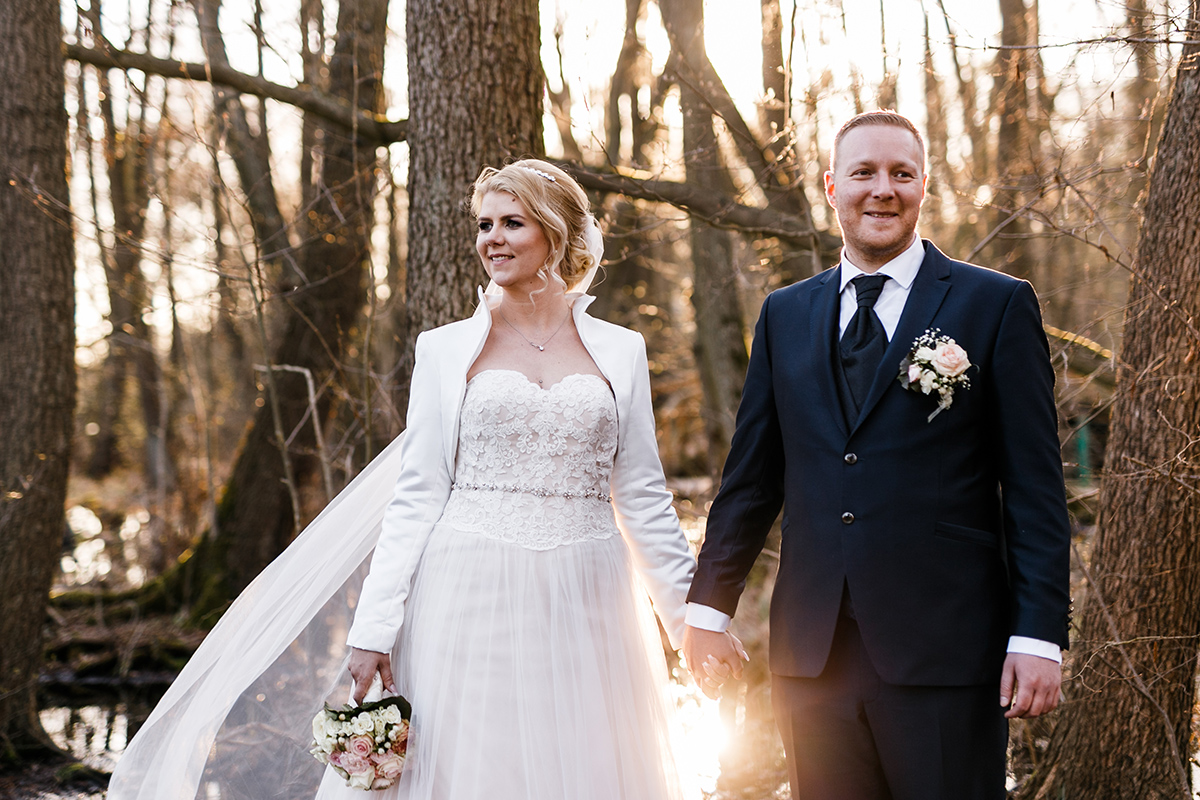 Hochzeitsoutfit Winter für Braut aus Prinzessinen Hochzeitskleid mit Tüll und Spitze und kurzer weißer Jacke oder Bolero und Bräutigam in schwarzem Hochzeitsanzug bei Fotoshooting im Wald - Hochzeitsfotografin aus Berlin hat Winterhochzeit am Wasser in Brandenburg im Landgasthof "Zum Mühlenteich" begleitet © www.hochzeitslicht.de