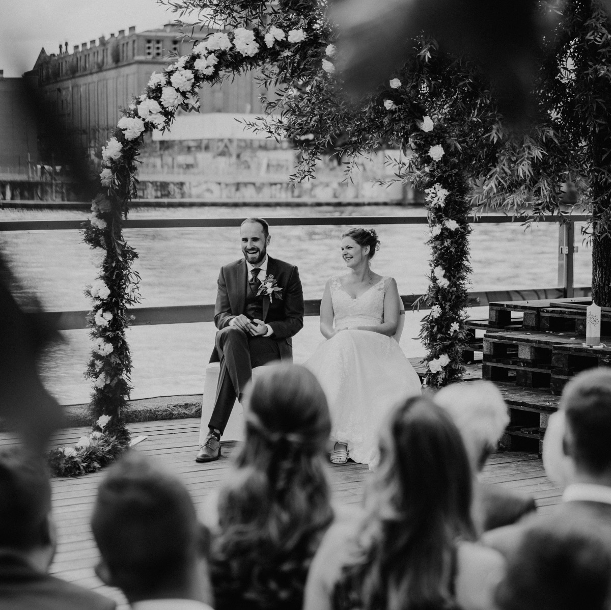 authentisches Hochzeitsfoto von Brautpaar unter Traubogen mit weißen Blumen auf Terrasse am Spreeufer - 89 Lighthouse White Spreelounge Berlin Hochzeitsfotograf © www.hochzeitslicht.de