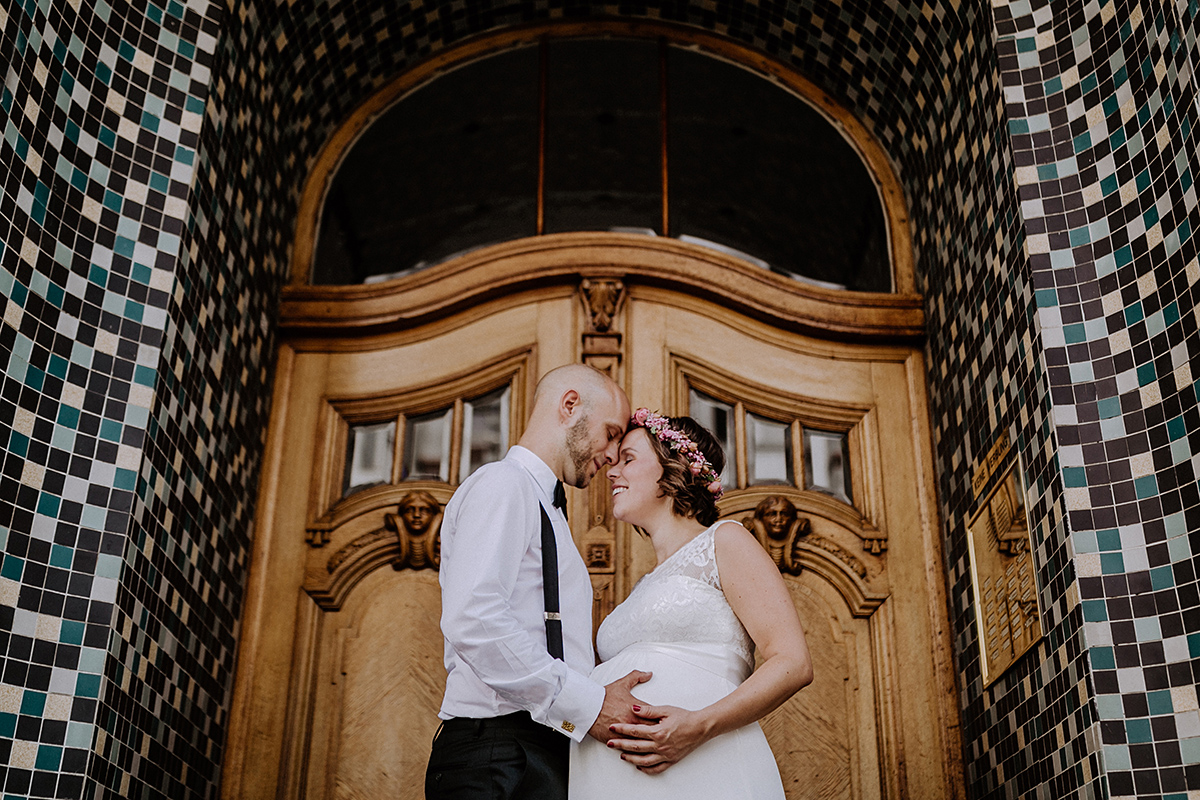 Hochzeitsfoto Brautpaar schwanger heiraten Standesamt im Sommer, Braut mit Blumenkranz und Kurzarm Brautkleid für Schwangere mit Spitzenoberteil und Gürtel, Bräutigam in schwarzem Anzug mit Hosenträgern und Fliege - Hochzeitsfotograf Standesamt Rathaus Schöneberg © www.hochzeitslicht.de