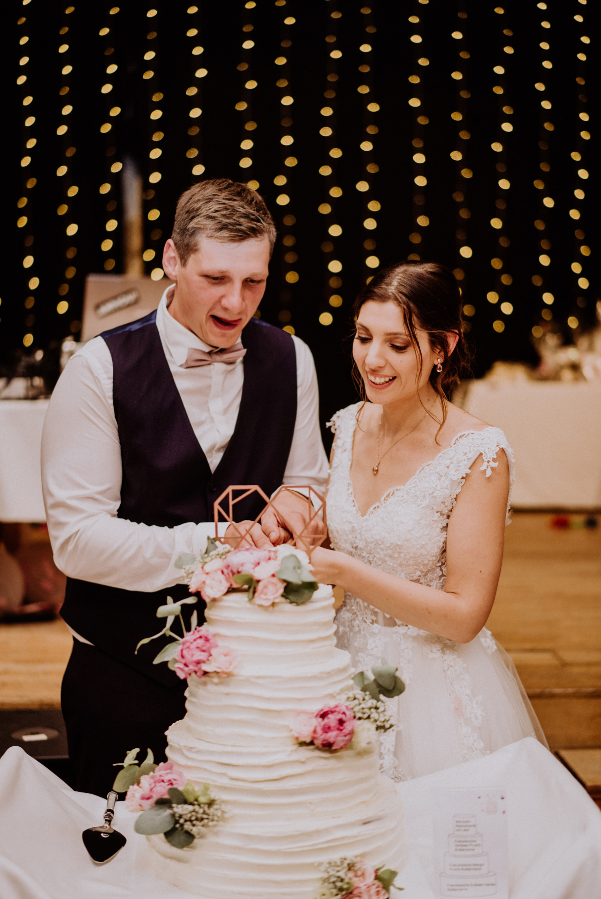 Hochzeitsfoto Hochzeitstortenanschnitt mit Lichterketten Deko Scheune; vierstöckige weiße Hochzeitstorte mit roségoldenem Herztopper, Pfingstrosen - Scheunenhochzeit in Brandenburg - freie Trauung Kulturscheune Thyrow von Hochzeitsfotograf Berlin © www.hochzeitslicht.de #hochzeitslicht