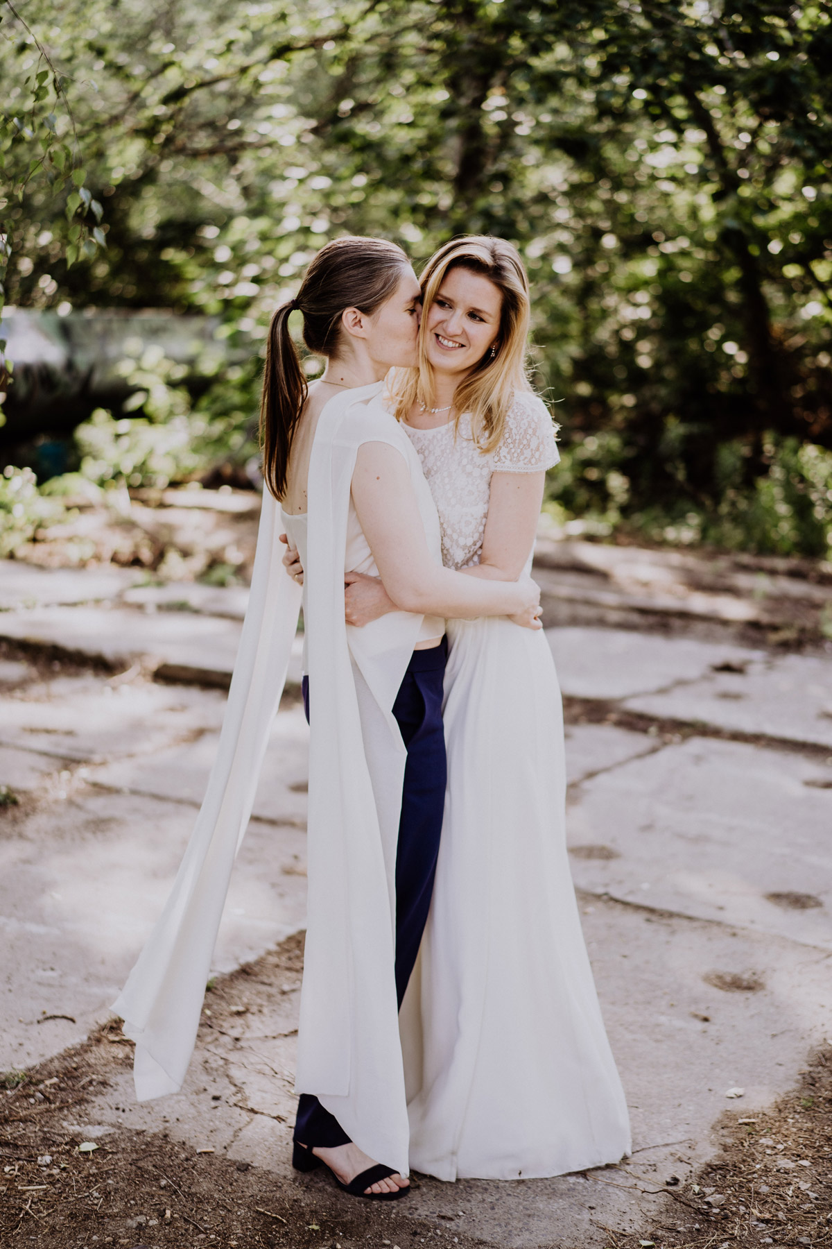 lesbische Hochzeit: Paarshooting Idee Natur nach Standesamt homosexuelle Frauen küssen sich im Fotoshooting auf Wange; gleichgeschlechtliches Hochzeitsbild von Hochzeitsfotografin aus Berlin © www.hochzeitslicht.de #hochzeitslicht; Outfit Bräute Hochzeitskleid Zweiteiler mit schlichtem, fließenden A-Linien-Rock und Oberteil aus Spitze und als Alternative zum Brautkleid dunkle Hose und weiße Seiden-Bluse mit offenen Schleierärmeln #homo #hochzeit #gaywedding #gay #wedding #photoshooting #brides