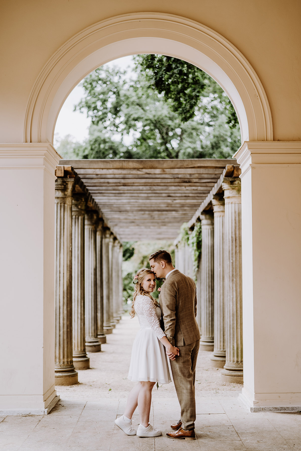 Hochzeitsfotos Idee nach standesamtlicher Hochzeit in Berlin Pankow von Hochzeitsfotograf Berlin © www.hochzeitslicht.de #hochzeitslicht im Park vor Säulen - Braut trägt weiße Turnschuhe zum kurzen Standesamt Kleid mit langen Ärmeln aus Spitze