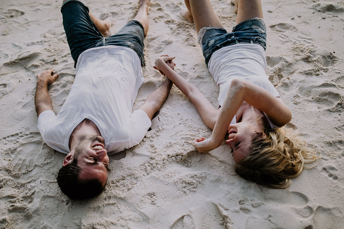Seychelles photographer takes photo of laughing couple lying in the sand at pre-wedding photoshoot